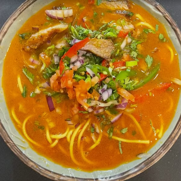 Spicy noodle soup with chicken, red onions, tomatoes, and cilantro in a light green bowl.