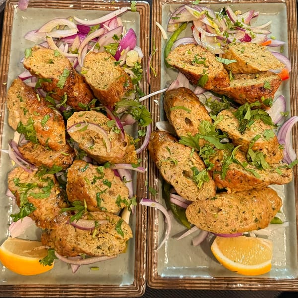 Two plates of sliced kebabs with red onion, green chilies, cilantro, and lemon wedges.