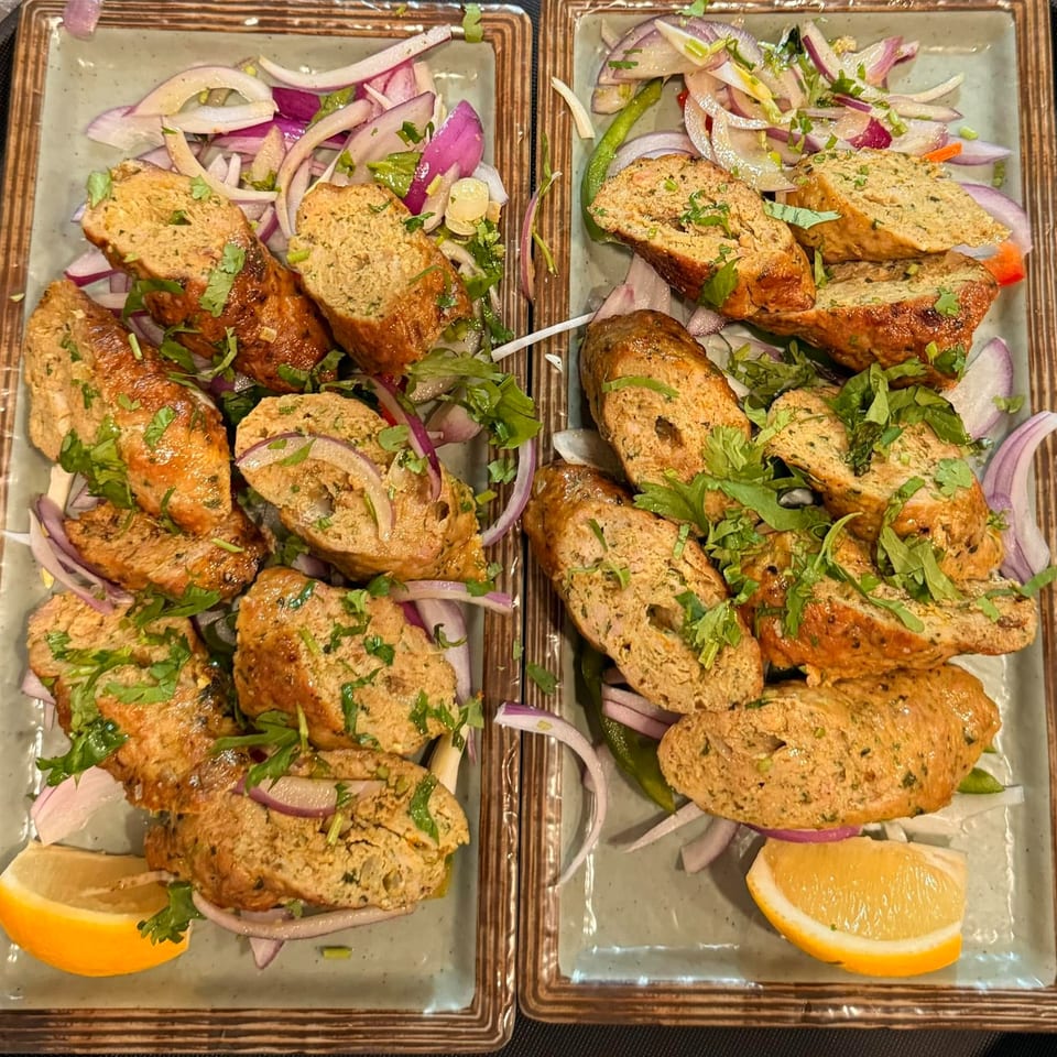 Two plates of sliced kebabs with red onion, green chilies, cilantro, and lemon wedges.
