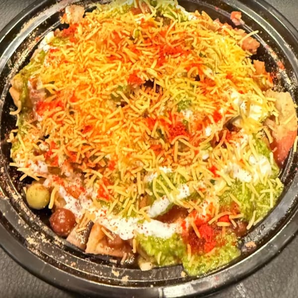Close-up of a colorful Indian chaat dish with crispy noodles, sauces, and spices in a black bowl.