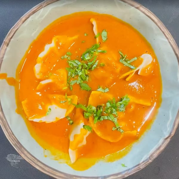 Dumplings in a vibrant orange sauce, garnished with fresh green herbs, served in a light blue bowl.