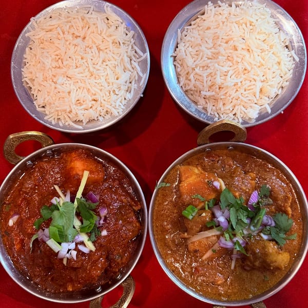 Two Indian curries in metal bowls with rice, garnished with herbs and onions, on a red table.