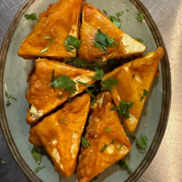 Six triangular pieces of fried paneer or tofu, coated in a reddish-orange sauce and garnished with cilantro.